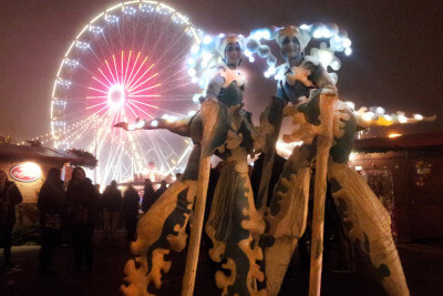 2 beleuchtete Stelzenläufer vor einem Riesenrad