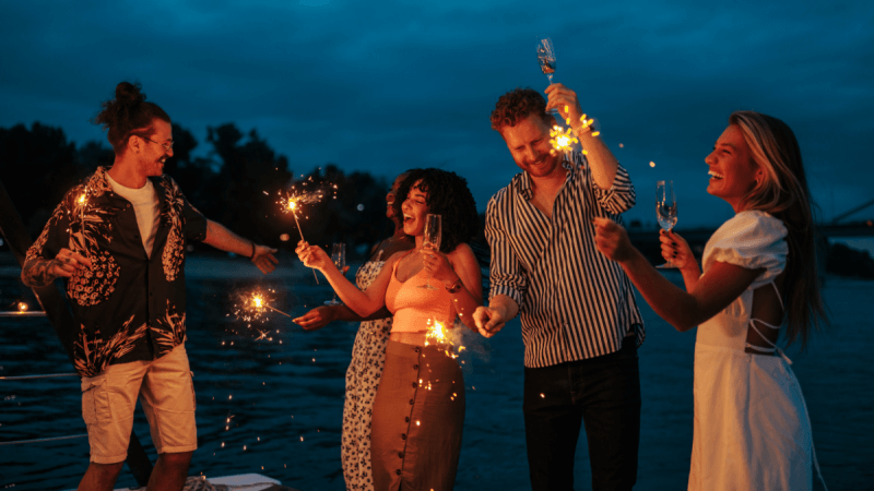 Fünf junge Menschen feien auf einem Boot mit Gläsern und Wunderkerzen in der Hand bei Abenddämmerung