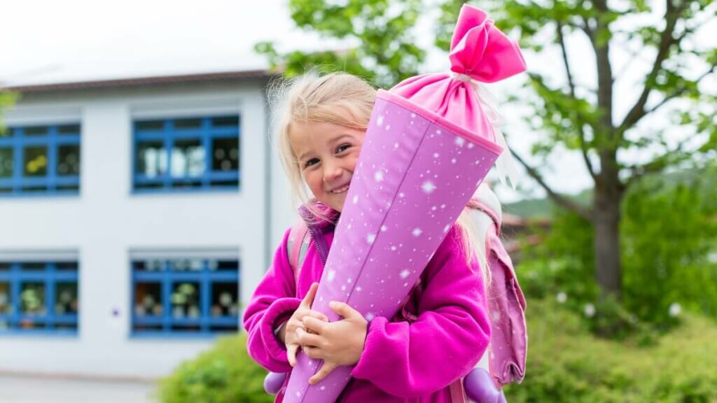 Lachendes Mädchen steht mit pinker Schultüte vor einem Schulgebäude