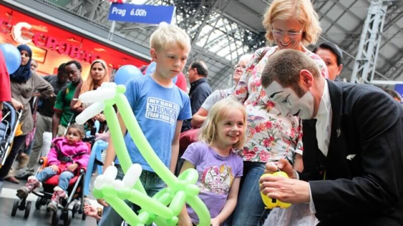 Mann mit weiß geschminktem Gesicht formt zwei Kindern Figuren aus Luftballons am Bahnhof