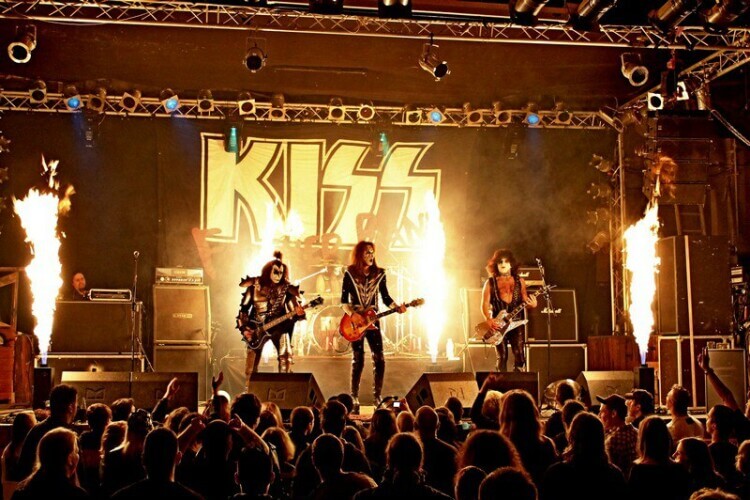 Rockmusiker der KISS Tribute Band stehen mit Gitarren auf der Bühne, dahinter schießen Feuerfontänen Ihre Flammen empor